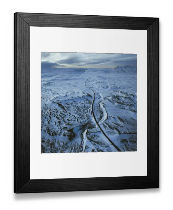 Framed Print : Aerial view of a long road winding through a vast snowy landscape with distant mountains under a cloudy sky Framed Print : Aerial view of a long road winding through a vast snowy landscape with distant mountains under a cloudy sky