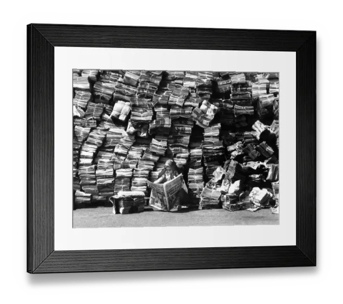 Framed Print : Woman reads newspaper in front of a big pile of newspapers