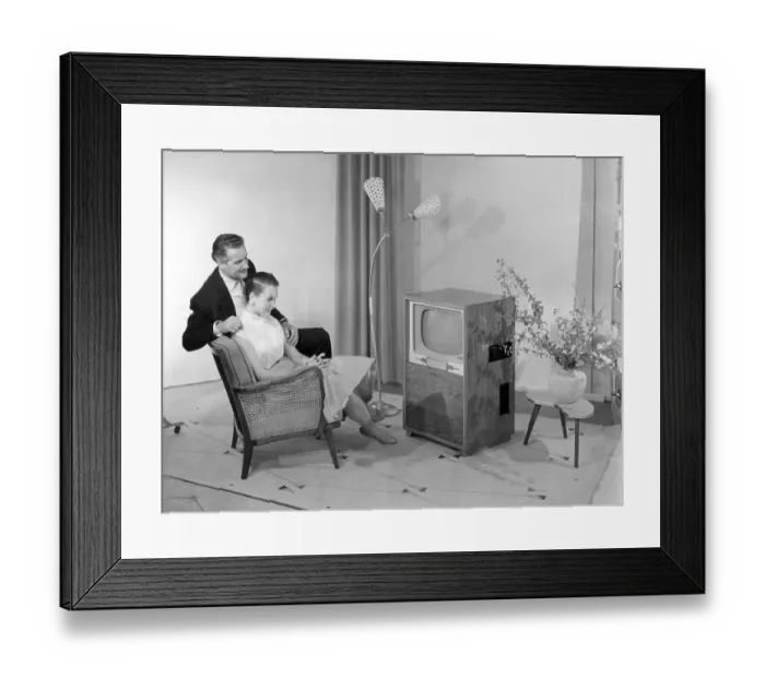Framed Print : Man and woman sitting on an arm chair in front of a television