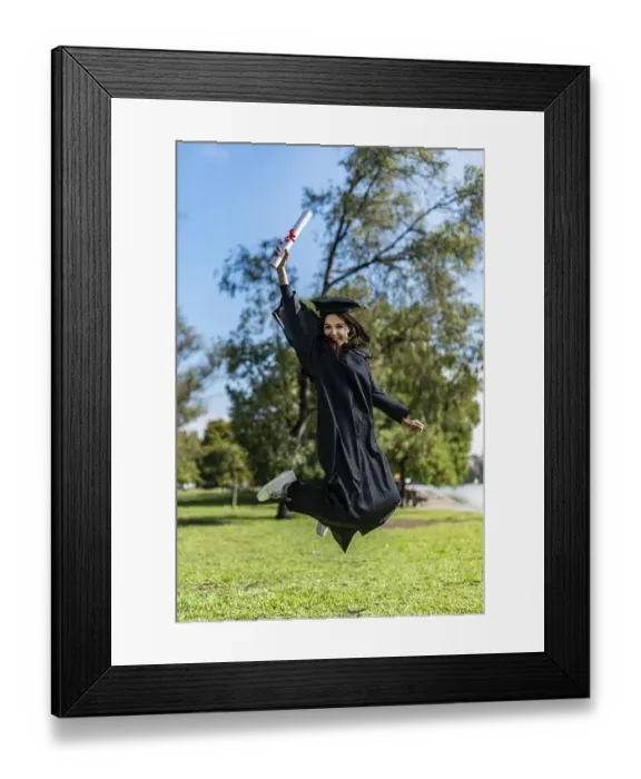 Framed Print : Happy graduated girl in a black gown jumping while holding her diploma