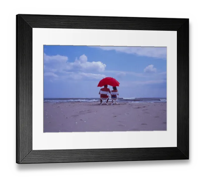 Framed Print : Couple on the beach, seated under red sunshade