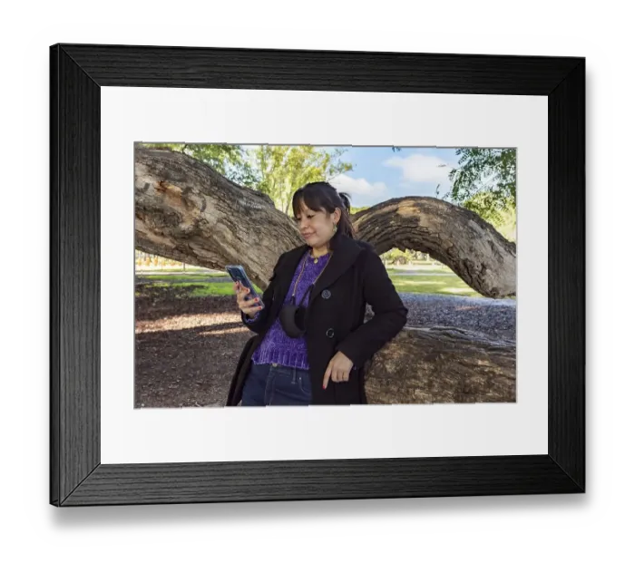 Framed Print : A woman is reading a text message on her mobile phone while leaning on a tree in the park