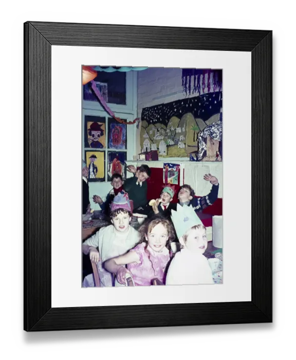 Framed Print : Christmas party time in primary school classroom children wearing hats and eating pulling funny faces, UK, c 1960