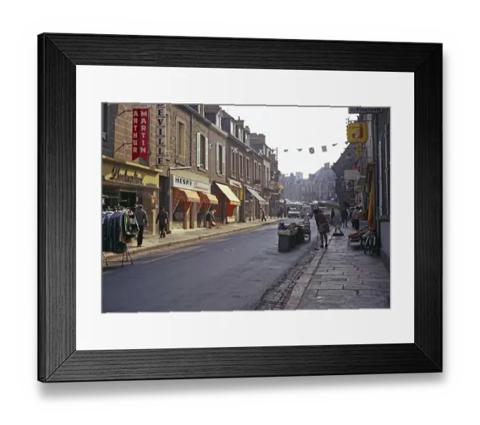 Framed Print : Eine Reihe von Geschaeften mit Menschen, die die zum Marktplatz fuehrende Strasse buersten, vermutlich in Quimper, Bretagne, Frankreich, 1974 Framed Print : Eine Reihe von Geschaeften mit Menschen, die die zum Marktplatz fuehrende Strasse buersten, vermutlich in Quimper, Bretagne, Frankreich, 1974