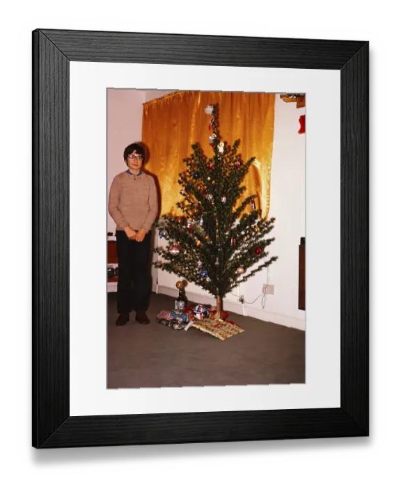 Framed Print : Junge steht vor einem kuenstlichen Weihnachtsbaum mit einem Geschenk darunter, Vorhaenge zugezogen, britische Kultur 1974