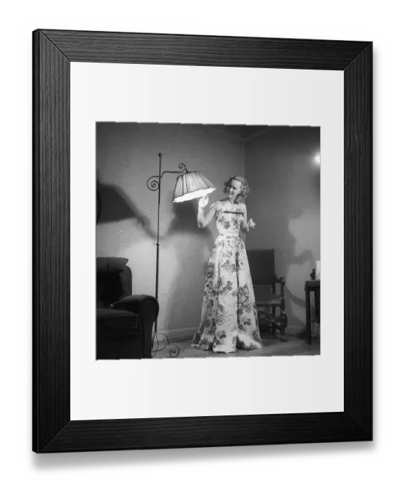 Framed Print : Fashion of 1946. A woman pictured wearing the typical fashion of the postwar era. She is wearing a long dress, the fabric of which is a pattern of flowers and leaves. She is standing in a room by a floor lamp which is lit. Kristoffersson ref Y129-2