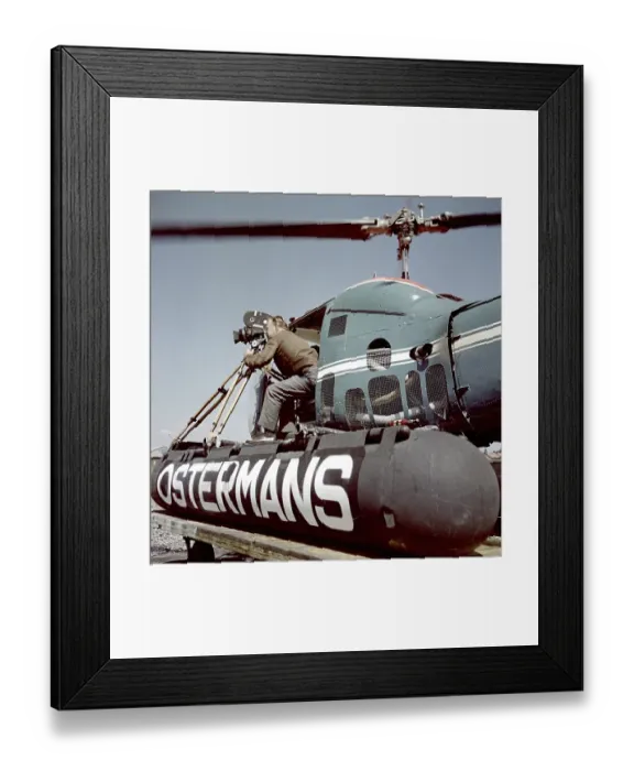 Framed Print : Filming in the 1950s. A cinematographer is standing half way out of a helicopter when filming something. The film camera is mounted on a tripod. Ostermans was a swedish company specialized in helicopter transports