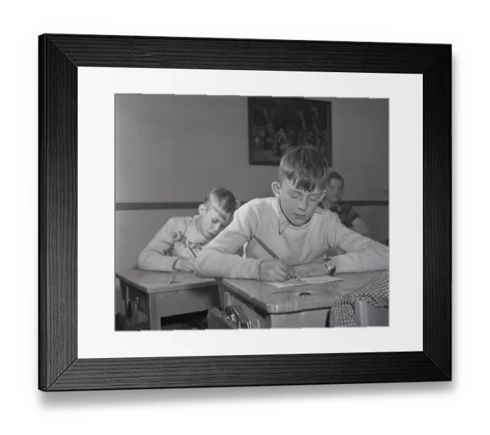 Framed Print : School in the 1940s. The interior of a classroom with a class of children sitting neatly at their desks. A boy is sitting at the front of the class, writing with an ink pen. The school desks had a lid that opened