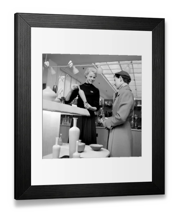 Framed Print : Shopping in the 1950s. A young woman in a store where a sales woman is presenting some of the stores selection of fine cermaics and decorative items designed by the famous designer Stig Lindberg Framed Print : Shopping in the 1950s. A young woman in a store where a sales woman is presenting some of the stores selection of fine cermaics and decorative items designed by the famous designer Stig Lindberg
