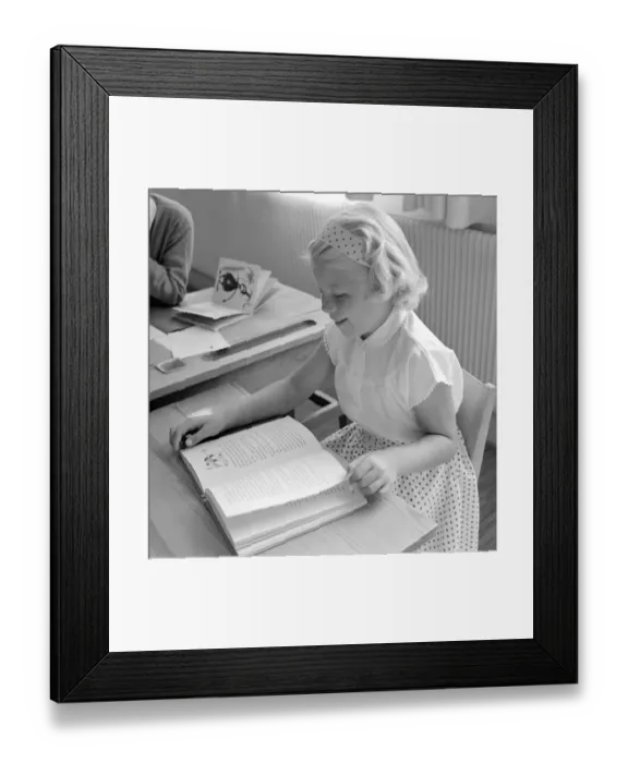 Framed Print : School in the 1950s. A girl is seen sitting at her desk in the classroom reading from the schoolbook in front of her
