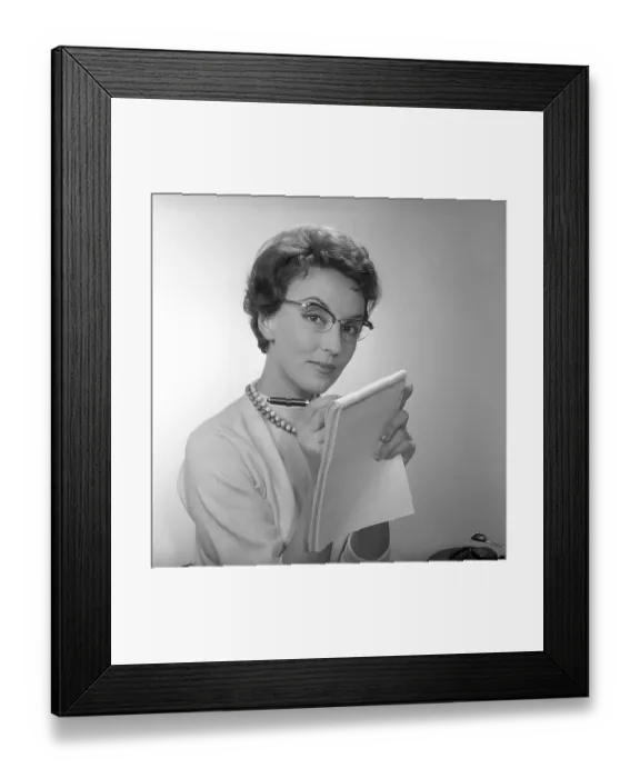 Framed Print : In the 1950s. A woman at a desk with a typewriter visible holds a notepad and writes in it with a pen