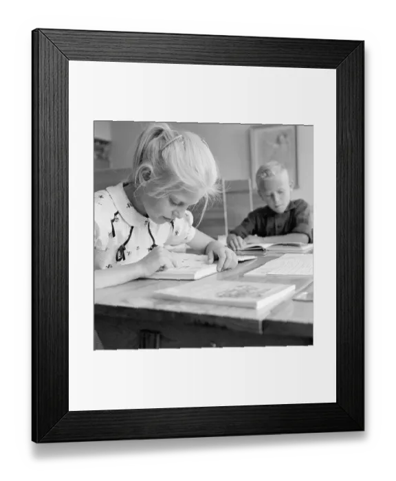 Framed Print : School in the 1950s. A girl is seen sitting at her desk in the classroom reading from the schoolbook in front of her