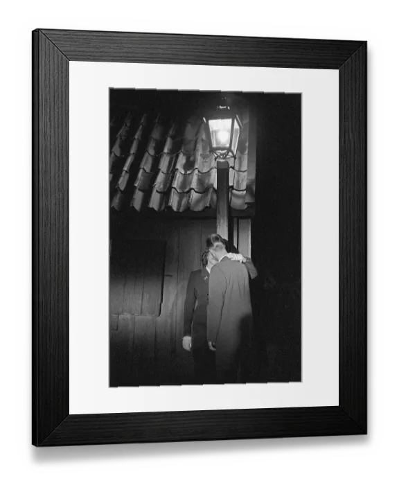 Framed Print : Love in the 1950s. A young couple in the evening are standing under a street lamp kissing
