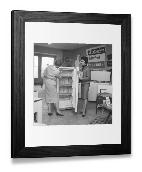 Framed Print : Freeze in the 1960s. A young woman works as a salesperson in an appliance store. She shows a customer an ITT brand refrigerator. Deep-frozen food would be the start of a revolution in household habits and food storage