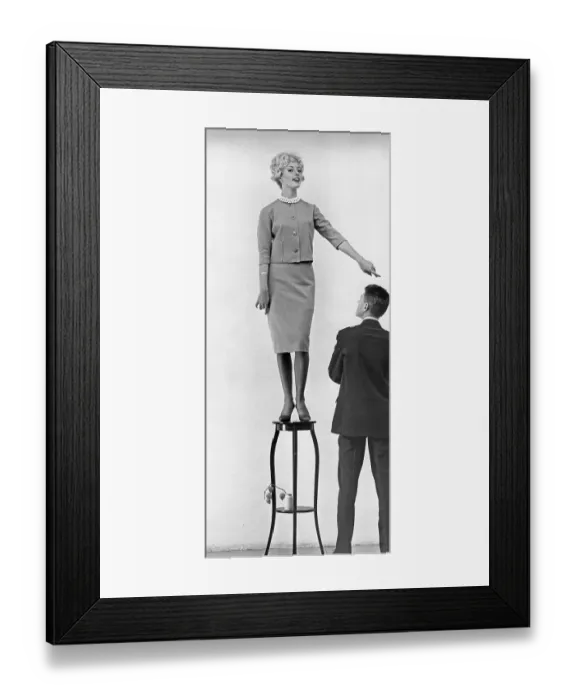 Framed Print : In the 1950s. A young woman seen standing on a piece of furniture higher than the man standing next to her Framed Print : In the 1950s. A young woman seen standing on a piece of furniture higher than the man standing next to her