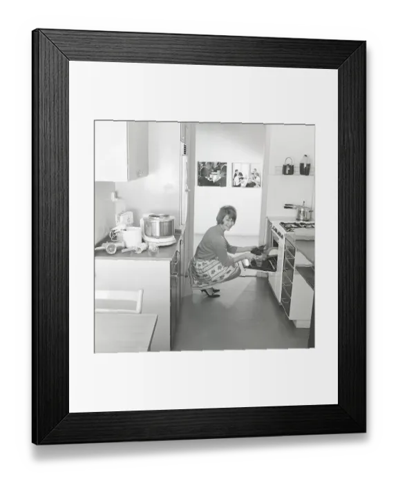 Framed Print : In the kitchen in the 1960s. A young woman in the kitchen taking out the ready baked breads from the oven