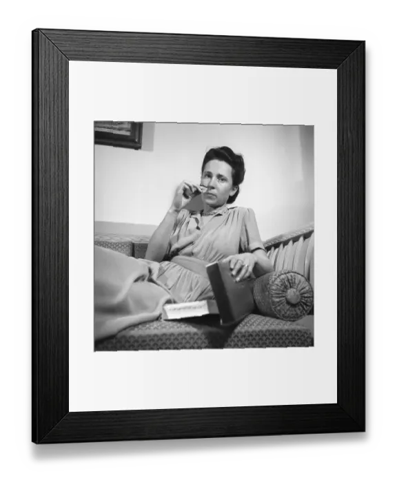 Framed Print : In the 1940s. A young woman is sitting on the couch with a piece of chocolate or candy in her hand from a box next to