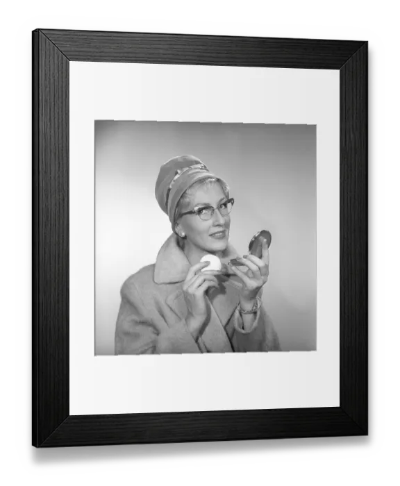 Framed Print : 1950s makeup. A woman is looking at herself in her pocket mirror and improves on her makeup. She is wearing a fashionable hat, typical 1950s glasses and a coat. 1958 Kristoffersson ref CB96-5