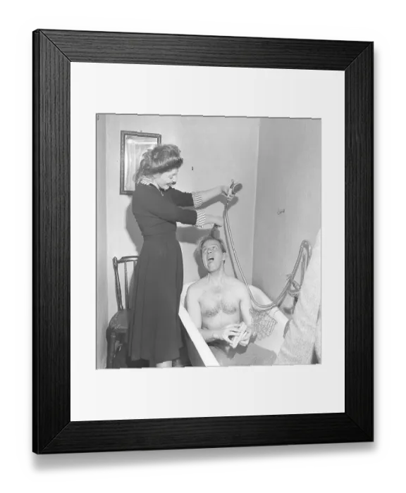 Framed Print : Couple in the bathroom in the 1940s. A man sits in a bathtub with only his pants on. A young woman laughs and playfully holds his hair and the shower head to pretend to shower him. Sweden 1949. Kristoffersson ref AT43-4