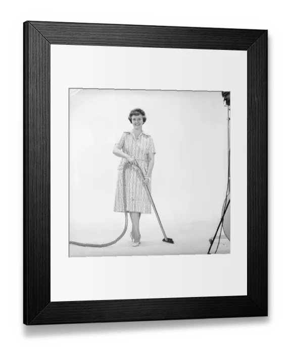 Framed Print : Cleaning day in the 1960s. A woman is standing holding a vacuum cleaner