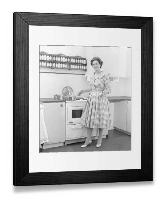 Framed Print : In the 1950s. A woman in a typical 1950s kitchen with wooden cabinets and a set of spices on shelfes above the cooker