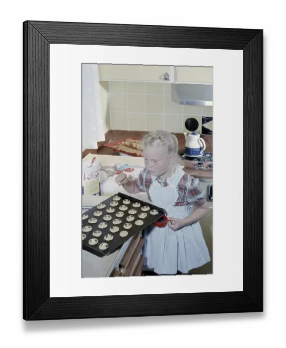 Framed Print : In the early 1960s. A girl nicely dressed in an apron puts raisins on top of the almost ready to bake cookies Framed Print : In the early 1960s. A girl nicely dressed in an apron puts raisins on top of the almost ready to bake cookies