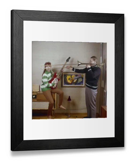 Framed Print : In the 1960s. A couple is seen playing the trombone and an electric guitar, looking happy and playful. To the left a radio gramophone. A combined music furniture that could play records and radio in the same unit. Often a large piece of furniture Framed Print : In the 1960s. A couple is seen playing the trombone and an electric guitar, looking happy and playful. To the left a radio gramophone. A combined music furniture that could play records and radio in the same unit. Often a large piece of furniture