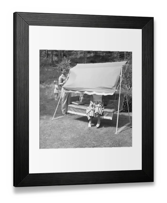 Framed Print : Summer in the 1950s.A woman is sitting in a hammock but her husband plays with her