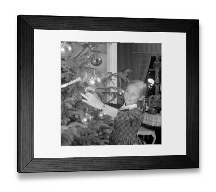 Framed Print : Christmas in the 1960s. A young girl is standing by the christmas tree decorating it Framed Print : Christmas in the 1960s. A young girl is standing by the christmas tree decorating it