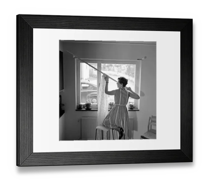 Framed Print : 1960s home decorating. A woman at home putting up the curtains in the room window. All by herself she balances on a chair and a stool to manage it. Sweden 1966