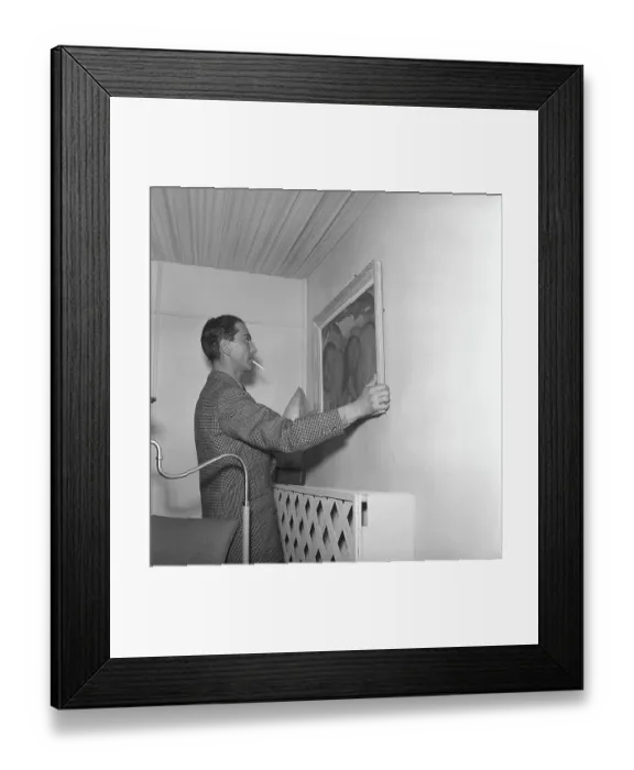 Framed Print : In the 1940s. A man tries to put a painting up on the wall and is carefully adjusting it so it hangs right Framed Print : In the 1940s. A man tries to put a painting up on the wall and is carefully adjusting it so it hangs right