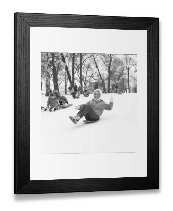 Framed Print : Winter in the 1960s. A young woman is sliding down a hill on a saucer shaped type plastic pulk