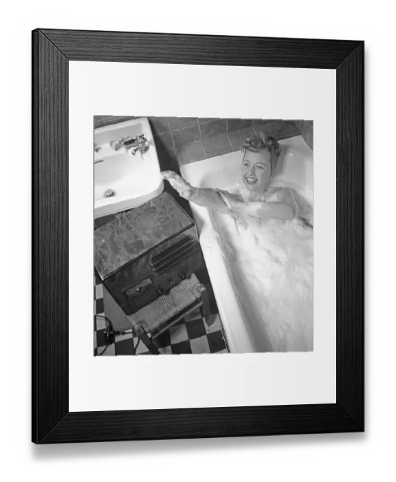 Framed Print : 1940s woman in the bathtub. A young woman is having a nice warm bath. She is careful about not getting her hair wet, and has it up under a scarf. A radio is standing on a chair beside the bathtub, and she is listening to it while bathing