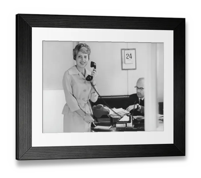 Framed Print : Office girl in the 1960s. A woman is standing in an office with a pen and notebook holding a telephone. A man is seen sitting at a desk. She is a secretary. Sweden 1961