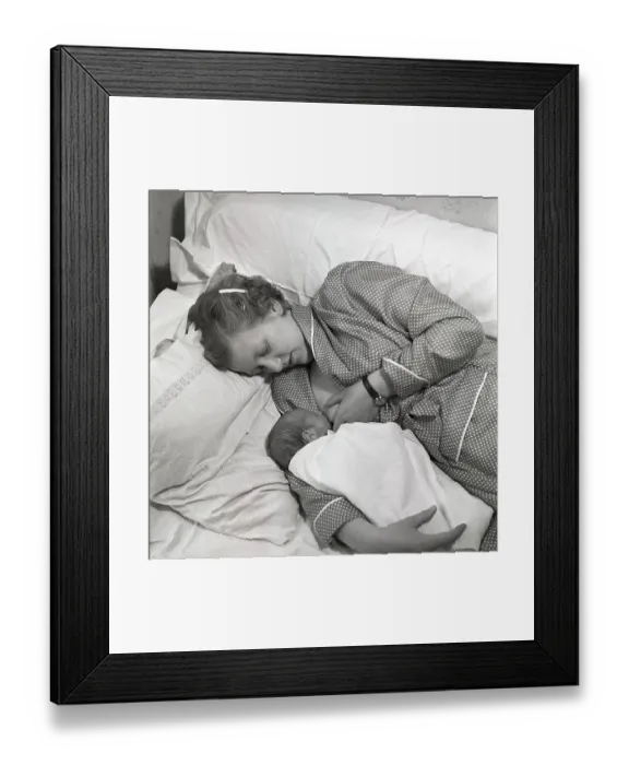 Framed Print : 1950s mother nursing her baby. A young woman breastfeeding her baby while lying in bed at home