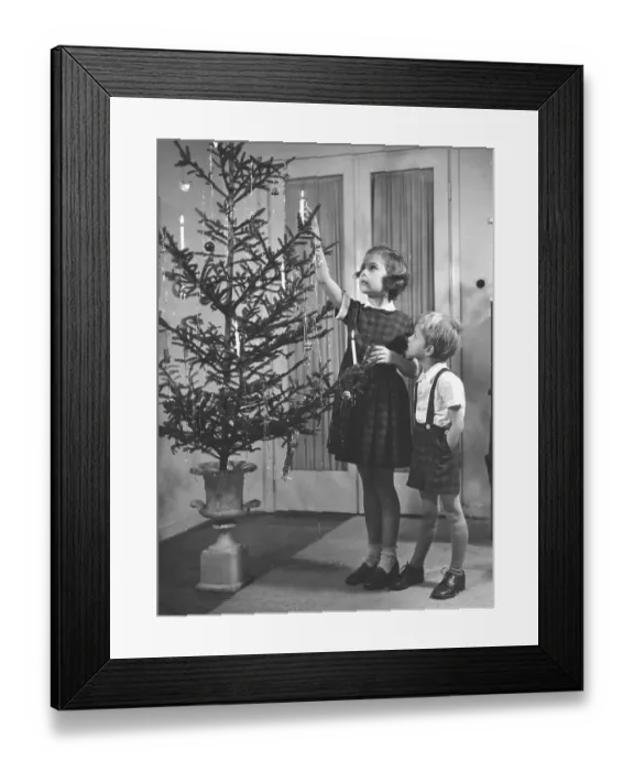 Framed Print : Christmas in the 1950s. A girl and a boy is lighting the candles in the christmas tree. They are nicely dressed for christmas. Sweden 1952