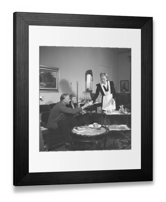 Framed Print : Living in the 1940s. A young woman working as a maid is serving tea and sandwiches to a man and woman sitting in the couch. She is neatly dressed in a white apron. Sweden 1946. Kristoffersson ref X18-3