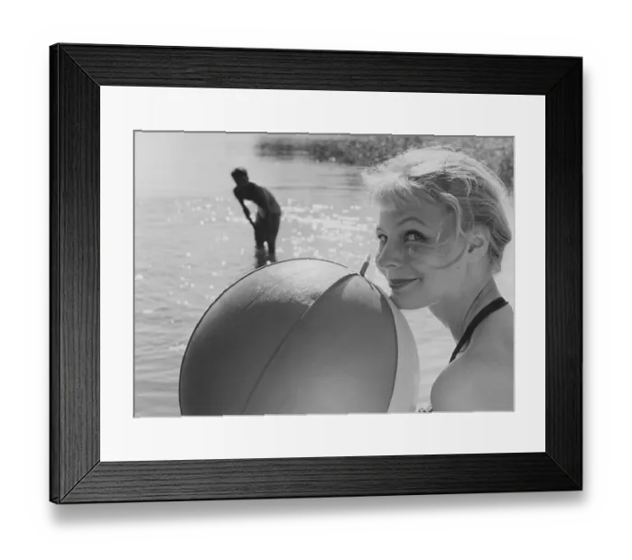 Framed Print : 1950s summer. A young blonde woman looks into the camera holding a typical ball to use when playing on the beach with