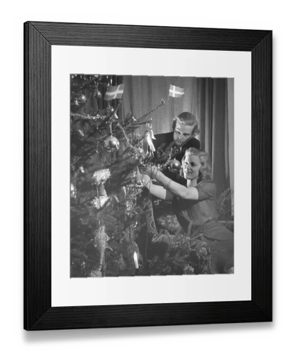 Framed Print : Christmas in the 1940s. A young couple is putting christmas decorations on the christmas tree Framed Print : Christmas in the 1940s. A young couple is putting christmas decorations on the christmas tree