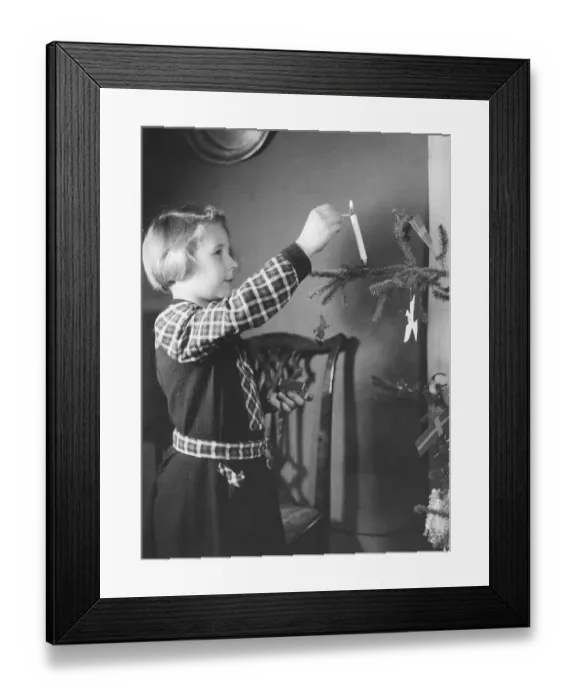 Framed Print : Christmas in the 1940s. A girl is decorating the christmas tree and is lighting a candle in the tree