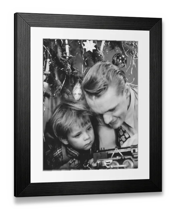 Framed Print : Christmas in the 1940s. A father and son are playing with a toy steam engine by the christmas tree
