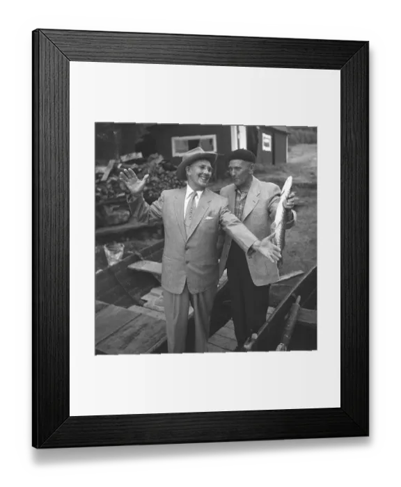 Framed Print : Fishing in the 1950s. A man has caught a fish and shows it up for his friend Framed Print : Fishing in the 1950s. A man has caught a fish and shows it up for his friend