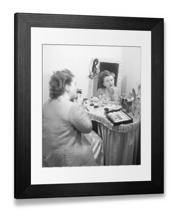 Framed Print : Cosmetics in the 1940s. A young woman at home is sitting at a make up desk and looking at herself in the mirror while putting on lipstick. There are many different kinds of cosmectics lying on the table in front of her. Sweden 1947