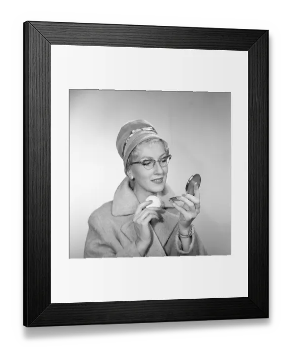 Framed Print : 1950s makeup. A woman is looking at herself in her pocket mirror and improves on her makeup