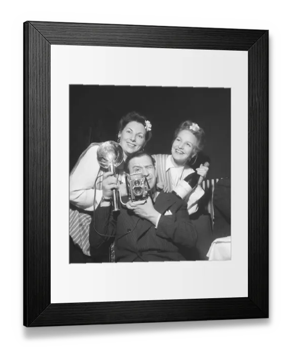 Framed Print : 1940s photographer. A man with a Rolleiflex camera and flash aims at something with two girls haning on his shoulders