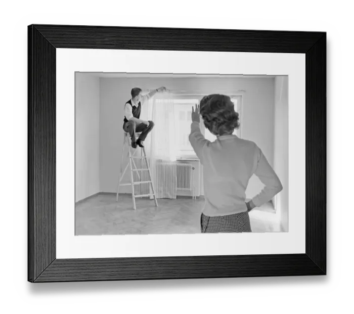 Framed Print : 1950s home decorating. A couple in their new apartment seen hanging the curtains