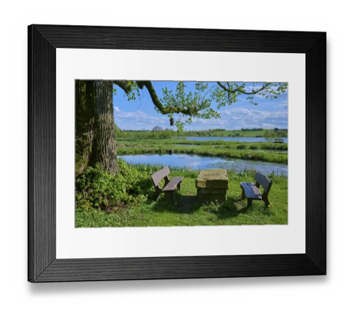 Framed Print : Ober- Moosersee surrounded by green fields and trees and a stone table with benches under a partly cloudy blue sky