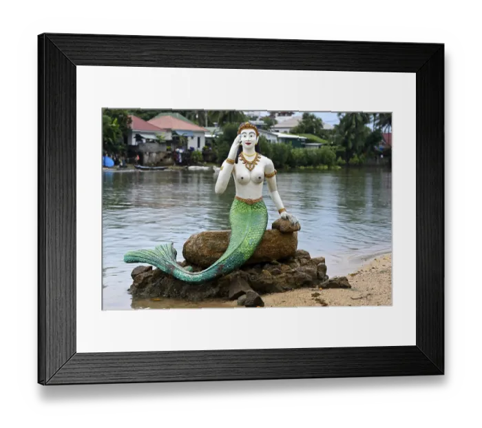 Framed Print : Greeting mermaid statue with white upper body and green tail fin sits on a stone on the shore