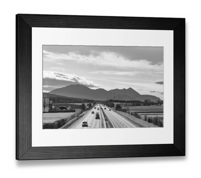 Framed Print : Busy motorway in front of a mountain backdrop in black and white with cloudy sky Framed Print : Busy motorway in front of a mountain backdrop in black and white with cloudy sky