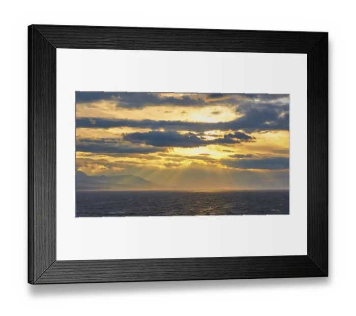 Framed Print : The sun sends its rays through the clouds over the sea near a mountain
